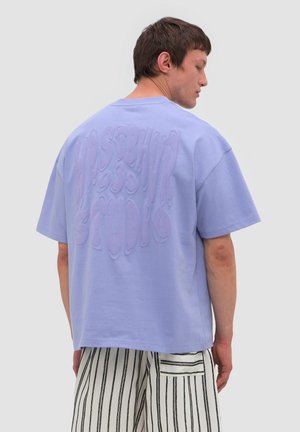 Jeune homme de dos portant un T-shirt lavande ample avec texte en relief et un pantalon blanc à rayures verticales noires avec une poche arrière.