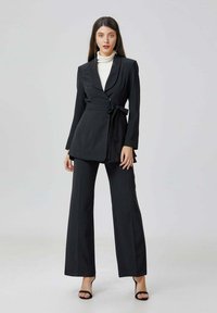 Femme portant un costume noir ajusté avec un pantalon large et un blazer ceinturé sur un col roulé blanc, se tenant contre un fond clair et uni.