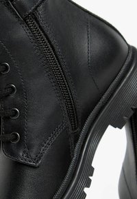 Botte de cheville en cuir noir avec un laçage à l'avant, une fermeture éclair sur le côté, une finition texturée et une semelle en caoutchouc épaisse.