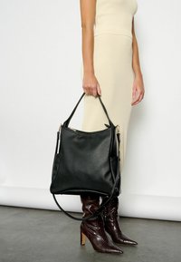 Čierna kožená hobo taška s textúrovaným povrchom, dvojitými uchami a zlatými kovovými doplnkami. Nosená s béžovými rebrovanými šatami a hnedými topánkami na opätku.