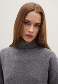 Giovane donna con capelli lisci castano chiaro che indossa un maglione grigio a collo alto, che guarda dritto in avanti con un'espressione neutra.