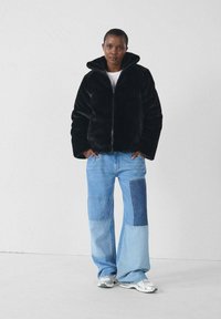 Giacca nera in pelliccia sintetica con zip e colletto alto, abbinata a pantaloni in denim a gamba larga con un motivo a patchwork in tonalità di blu chiaro e scuro.