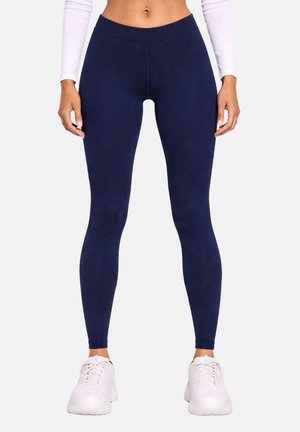 Femme portant des leggings bleu marine, un top court blanc à manches longues et des baskets blanches, debout avec les bras détendus le long du corps.