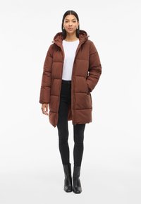 Giacca lunga marrone imbottita con cappuccio, caratterizzata da una chiusura con zip, grandi tasche e un design trapuntato, abbinata a jeans skinny neri e stivaletti.