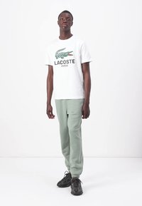 Witte katoenen t-shirt met een groen krokodillenlogo en tekst "LACOSTE PARIS", gecombineerd met zachte groene sweatpants en donkere sneakers.