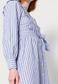 Sininen ja valkoinen raidallinen mekko, jossa on nappi etuosa, röyhelökaula-aukko, pitkät hihat ja vyötärölle sidottu vyö. Raikasta kangasta.