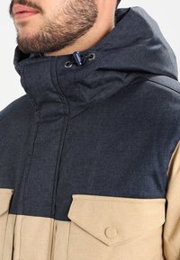 Chaleco acolchado que presenta una sección superior en azul marino y una sección inferior en marrón claro, con un alto cuello, capucha ajustable y bolsillos frontales.