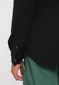 Maglietta nera a maniche lunghe con polsini abbottonati, tessuto strutturato e orlo curvo, abbinata a pantaloni verdi con un piccolo logo.
