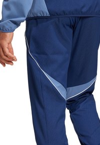 Marineblauwe trainingsbroek met een lichtblauwe accent en een witte zijstreep. Gemaakt van een gladde, lichte stof. Zichtbare hand aan de zijkant.