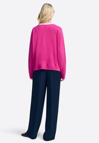 Blonde nainen seisoo, päällään kirkkaan pinkki pitkähihaisen neuleen ja tummansiniset leveälahkeiset housut, nähtynä takaa valkoista taustaa vasten.