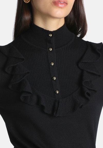 Femme portant un pull noir côtelé avec des boutons dorés sur le devant et des détails de tissu froncé près de l'encolure et des épaules.