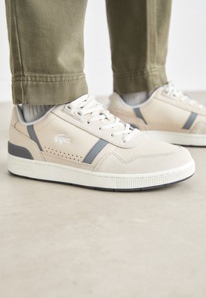 Beżowo-szare niskie sneakersy ze sznurówkami w kolorze białym, noszone z jasnoszarymi skarpetkami i spodenkami khaki, stojące na gładkiej powierzchni.