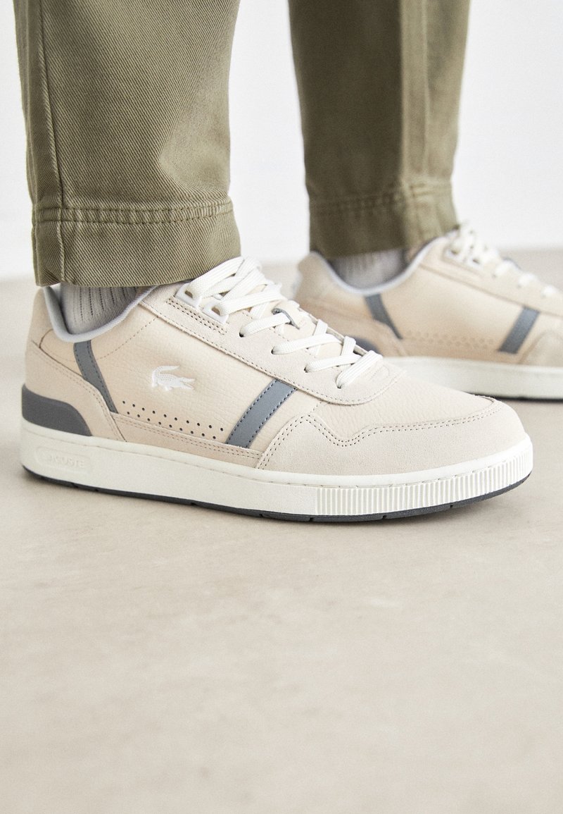 Baskets basses beige et grises avec des lacets blancs, portées avec des chaussettes gris clair et un pantalon kaki, debout sur une surface lisse.