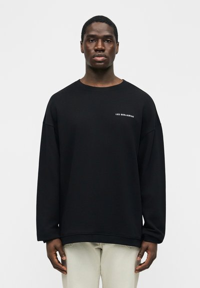 Άνδρας που φοράει μαύρο μακρυμάνικο φούτερ με το κείμενο «LES BENJAMINS» στο στήθος, συνδυασμένο με ανοιχτό μπεζ παντελόνι, στέκεται μπροστά σε απλό φόντο.