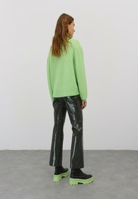 Grüner Strickpullover, glänzende schwarze Vinylhose und klobige schwarze Stiefel mit grünen Sohlen. Rückansicht, die Texturen und Farbkontraste zeigt.