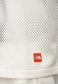 Tissu en maille blanche avec de petits trous et une étiquette rectangulaire rouge du logo North Face cousue près du bord inférieur.