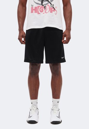 Mężczyzna w czarnych szortach Nike, białej koszulce z nadrukiem, białych skarpetkach Nike i białych butach sportowych Nike stoi na jednolitym tle.