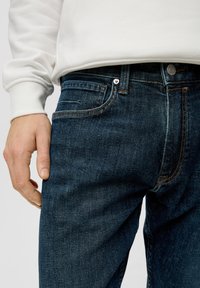 Jean en denim bleu foncé avec une coupe classique, avec des coutures orange, un bouton argenté et un design à cinq poches. Main reposant sur le côté.