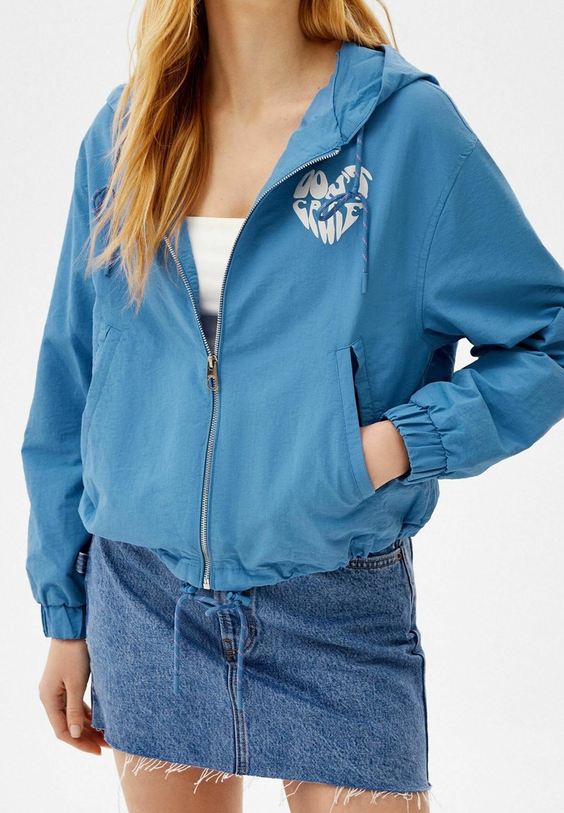Femme portant un sweat à capuche bleu zippé avec un logo circulaire sur un haut blanc et une jupe en jean bleu effilochée, main dans la poche.