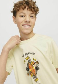 T-shirt giallo chiaro con una grafica di un re con corona, che tiene dei fiori. Il design include colori vivaci e un testo sopra l'immagine.