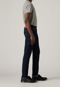 Camiseta gris de manga corta, jeans azul oscuro con costuras naranjas y zapatos negros con cordones. Los jeans tienen una parche de cuero marrón claro en la parte posterior.