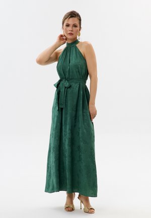 Donna con maxi abito verde scuro senza maniche a collo halter con motivo floreale e cintura in vita annodata, che indossa sandali beige con tacco alto.