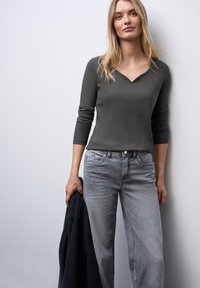 Žena s dlouhými blond vlasy, oblečená v těsném tmavě šedém ribbovaném topu a světle šedých džínách, držící černou bundu, stojící proti světlé zdi.