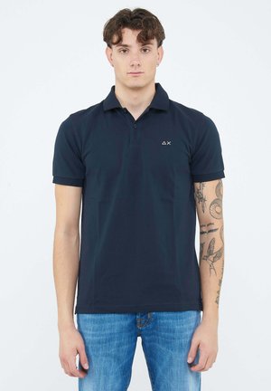 Giovane con capelli ricci che indossa una polo blu scuro e jeans blu, mostrando tatuaggi sul braccio sinistro su uno sfondo semplice.