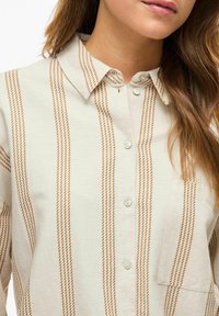Camisa de un beige claro con rayas verticales marrones, cuello tipo botón y bolsillo de parche. Confeccionada en una tela texturizada con un acabado suave.