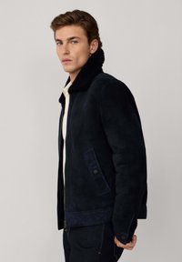 Marineblauwe suède jas met een zachte zwarte schapenvacht kraag. Voorzien van ritssluiting, zijzakken en een gestructureerde pasvorm. Effen textuur.