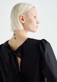 Abito nero con maniche a sbuffo, schiena scoperta e dettaglio a fiocco al collo. Tessuto liscio. Tatuaggi visibili sulla nuca.