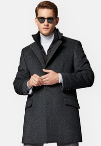 Ciemnoszara kurtka wełniana z klapami, z zamknięciem na guziki z przodu oraz dwoma bocznymi kieszeniami. Nosi się ją na jasnym golfie i zestawia z okularami przeciwsłonecznymi.