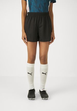 Persona con pantalones cortos deportivos negros, calcetines blancos Puma hasta la rodilla, botas de fútbol negras y una camiseta deportiva azul.