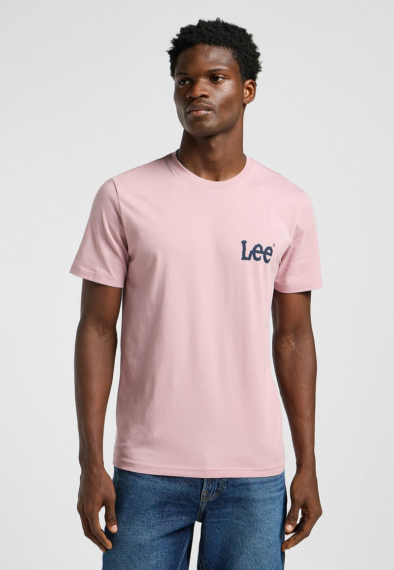 T-shirt di cotone rosa chiaro con collo rotondo, maniche corte e logo "Lee" blu sul petto sinistro. Indossato con jeans blu.
