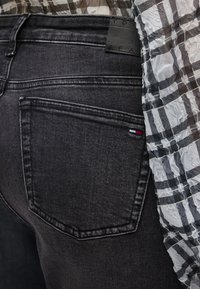 Κοντινό πλάνο πίσω τσέπης μαύρου τζιν με μικρό κόκκινο, λευκό και μπλε λογότυπο, σε συνδυασμό με τοπ από διαφανές ύφασμα με ασπρόμαυρο καρό σχέδιο.