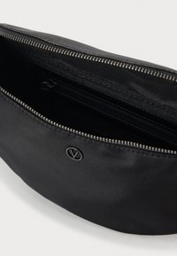 Črna torbica z zadrgo z majhnim okroglim kovinskim logotipom spredaj, pri kateri je delno odprt glavni predal z notranjim žepom z zadrgo.