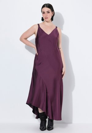 Vestito lungo con scollatura a V in satin viola scuro con spalline sottili. La silhouette drappeggiata presenta un orlo asimmetrico alto-basso e una consistenza liscia, completata da stivali neri eleganti.