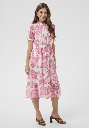 Žena sa usmieva, má na sebe biele midi šaty s ružovým vzorom paisley, krátkym rukávom, zapínaním na gombíky vpredu a hnedými sandálmi na podpätku.