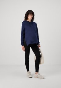 Sudadera azul marino con cordón, corte relajado y dobladillo trasero más largo; combinada con leggings negros y zapatillas deportivas beige. Bolsa blanca minimalista.