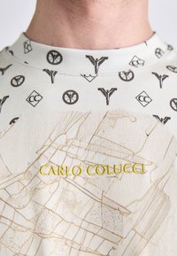 Lysfarvet stof med et grafisk design med små sorte mønstre og en broderet "CARLO COLUCCI" i gul. Teksturen er glat.