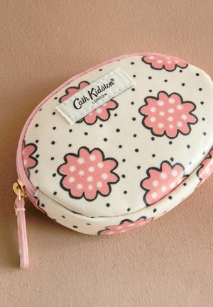 Krožna kosmetična torba z bež ozadjem, rožnatim cvetličnim vzorcem, črnimi pikami in roza zadrgo. Na strani je oznaka z logotipom.