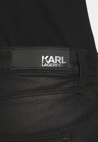 Los vaqueros de denim negro presentan una textura suave, costuras en contraste y un parche de cuero negro mate con texto en relieve.