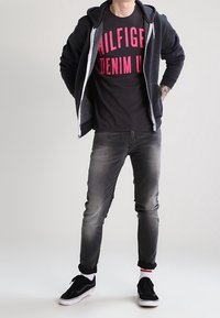 Neformální outfit zahrnující tmavě šedou mikinu s bílým zipem, černé tričko s jasně růžovým nápisem, vybledlé šedé džíny a černé tenisky.