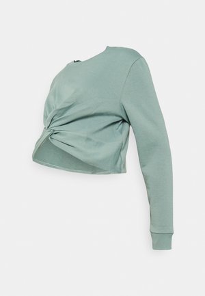 Chemise courte à manches longues vert clair avec un détail de nœud torsadé à l’avant, présentée sur un fond uni.