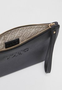 Pochette en cuir noir avec fermeture éclair, présentant un extérieur texturé, des garnitures dorées et une doublure intérieure à motifs. Logo visible à l'intérieur.