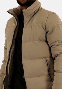 Beige pufferjacka med ribbad textur, hög krage, sidofickor och svart dragkedja. Fodret är av ett kontrasterande mjukt material.