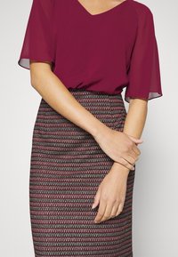 Femme portant un chemisier bordeaux à manches courtes et une jupe à motifs taille haute, les mains doucement croisées à la taille.