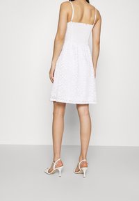White lace dress with a fitted bodice and flared skirt. Features thin straps and a textured pattern. Worn with white high-heeled sandals.