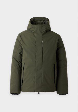 Un homme porte une veste à capuche vert foncé sur une chemise rose pâle, un jean bleu et des baskets blanches, sur un fond blanc uni.