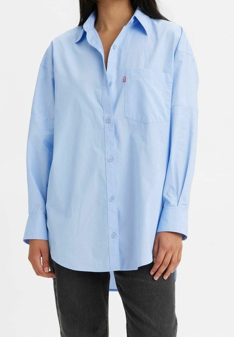 Femme portant une chemise bleu clair oversize à boutons avec une poche poitrine et un pantalon foncé, debout sur fond blanc.